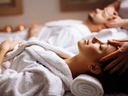 A couple in white robes laying down in a spa, enjoying head massages.